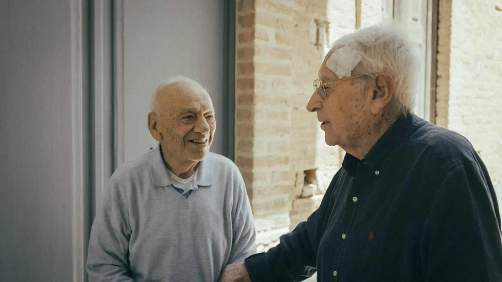 Due uomini anziani si salutano sorridendo davanti a una parete di mattoni al MuLab di Cannitello